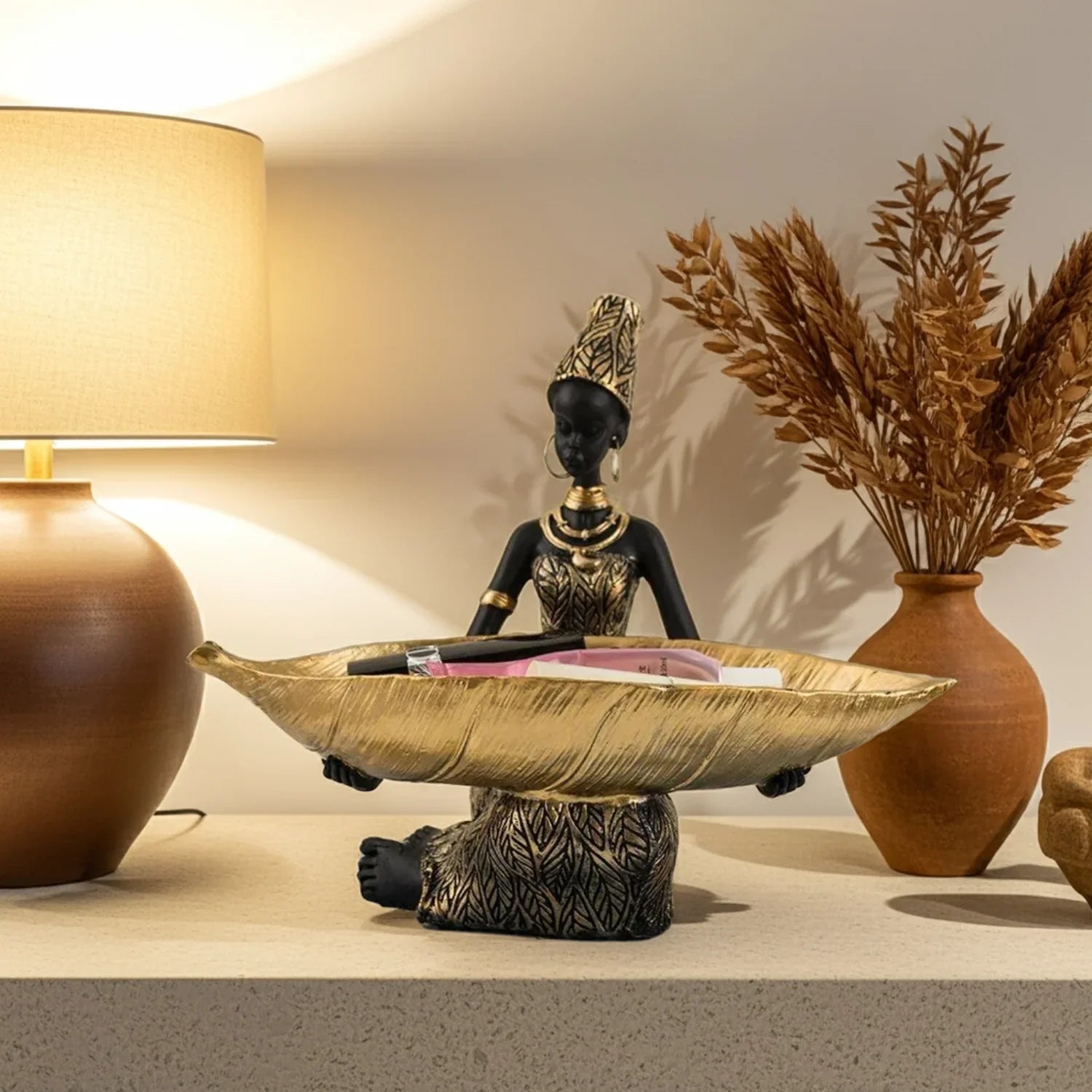 Decorative statue with a bowl on a table next to a lamp and vase.