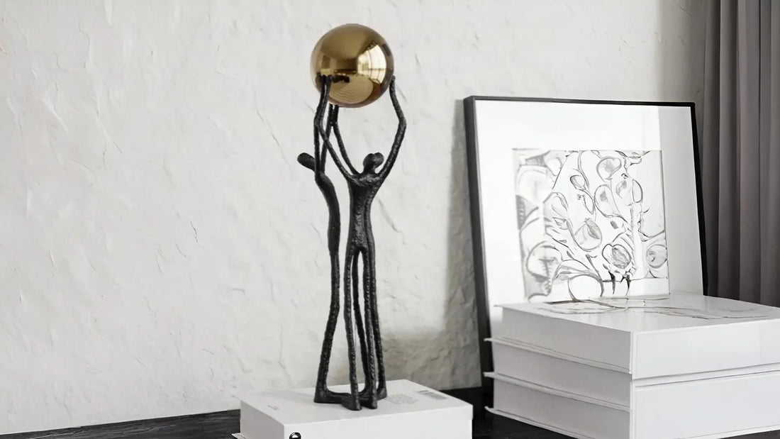 Abstract black metal sculpture of two figures lifting a golden sphere, displayed on stacked white books in a home decor setting