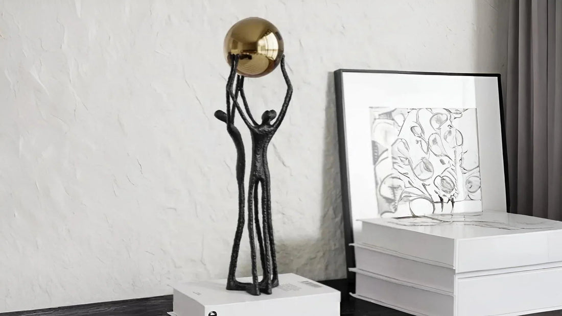 Black metal sculpture of two abstract figures holding a golden sphere on white books in modern room