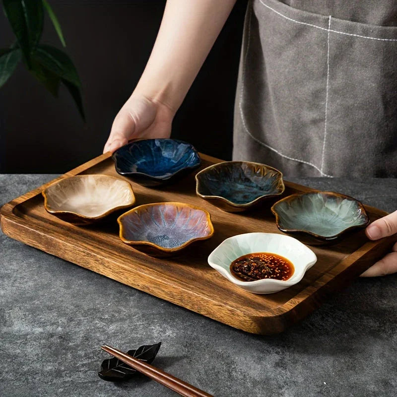 Hand holding wooden tray with six colorful ceramic dipping bowls, one containing sauce, on gray surface with chopsticks