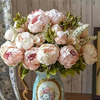 13 branches of realistic champagne peony silk flowers in a decorative vase with green foliage.
