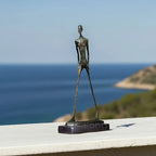 Bronze-colored abstract walking man sculpture with elongated limbs and textured metal surface on dark rectangular base