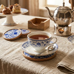Tea cup with blue floral patterns on a saucer, surrounded by a teapot, coasters, and pastries on a textured tablecloth.