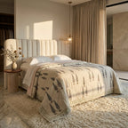 A styled bedroom scene featuring a bed dressed in a cream quilted coverlet with a gray tree pattern, set against a beige tufted headboard in a warm, sunlit room.