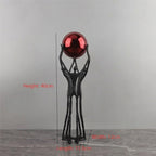 A black metal sculpture of two abstract figures holding a reflective red sphere, shown on a gray floor against a gray wall with overlaid dimension labels for height, width, and length.