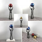 A collage showing a black figurative sculpture holding metallic decorative balls in different color options: red, blue, gold, and silver. The main view shows the black sculpture holding a red metallic ball, with other color variants displayed in inset panels on a gray marble-like surface against a light gray background.