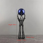 A decorative sculpture of three abstract human figures holding up a glossy dark blue sphere, shown on a gray surface against a gray wall. Red measurement text and lines indicate dimensions of the piece.