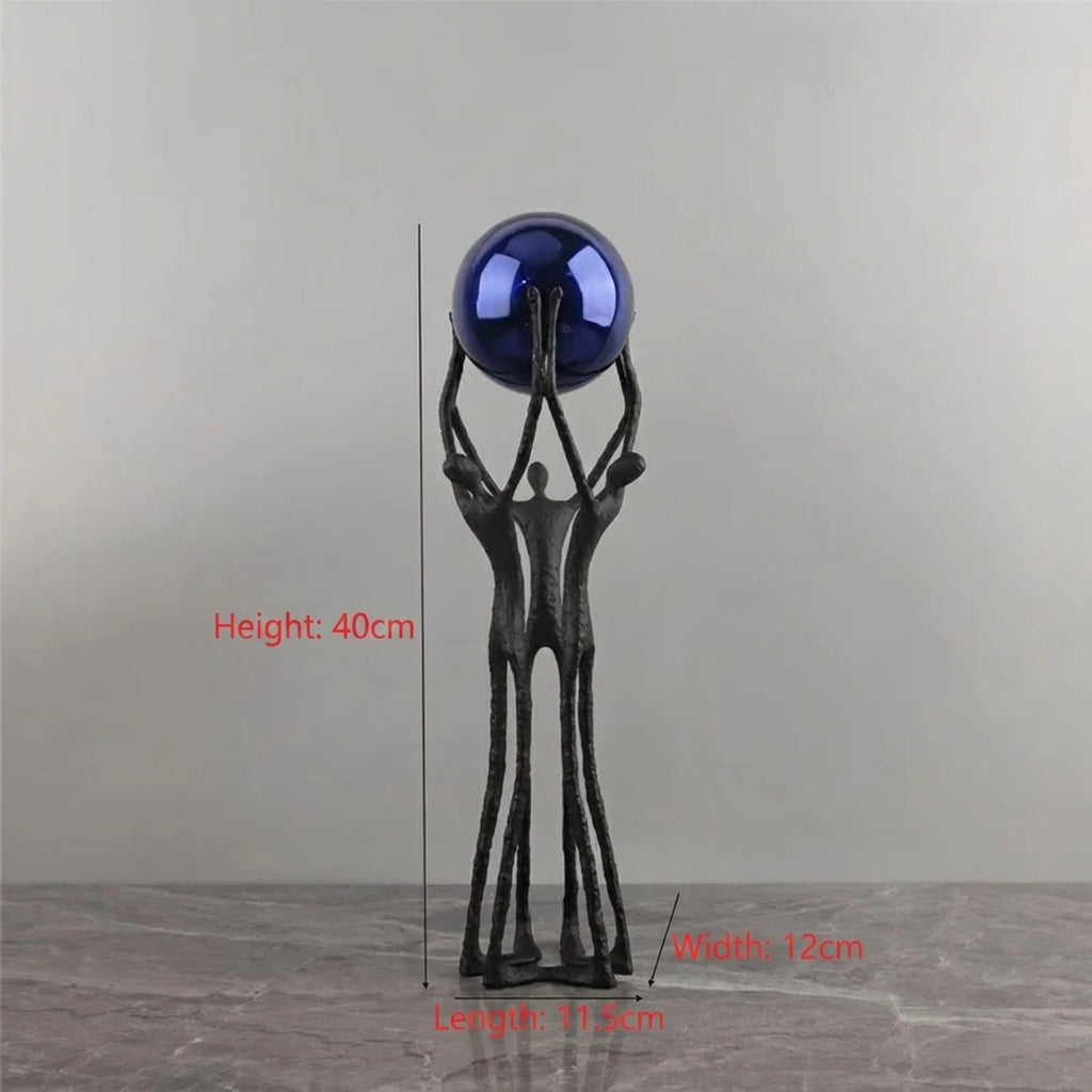 A decorative sculpture of three abstract human figures holding up a glossy dark blue sphere, shown on a gray surface against a gray wall. Red measurement text and lines indicate dimensions of the piece.