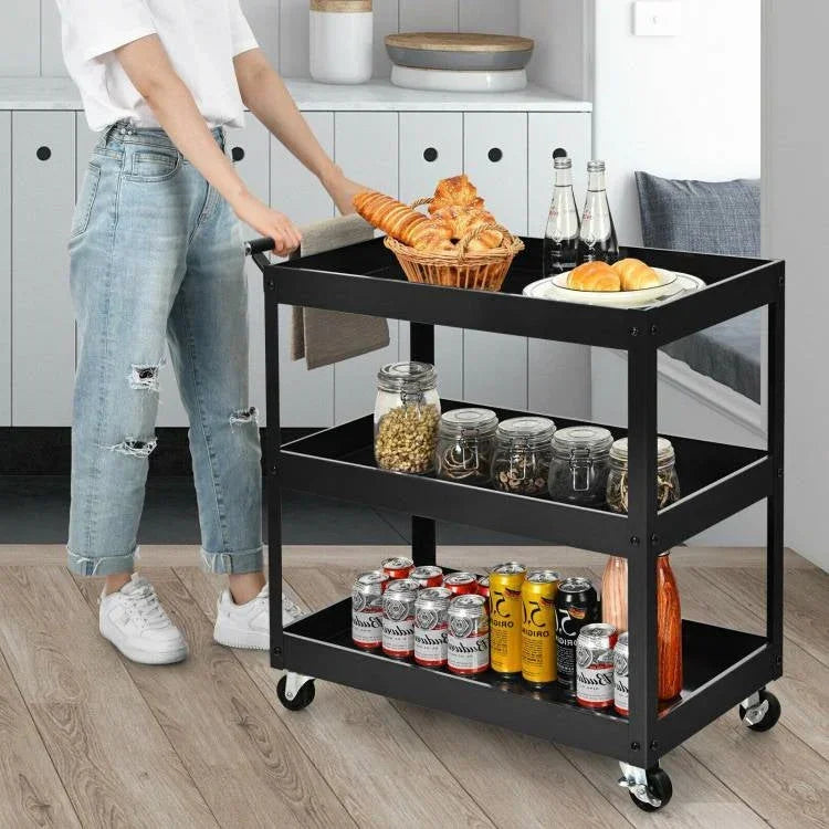 Person pushing a black three-tier cart filled with food and drinks in a kitchen.