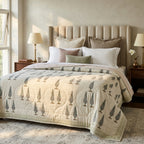 A neatly made bed in a softly lit bedroom, featuring a cream quilted comforter with a repeating tree motif in muted gray and taupe, layered with white and blush pillows against a beige tufted headboard.