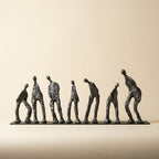 A set of seven dark metallic figurative sculptures of human figures in various poses, arranged in a row on a rectangular base against a warm beige background. [[SKU:HOA-SCU-012]]