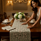 A woman sets a floral table runner on a dark wood dining table, surrounded by place settings, crystal glasses, and a silver floral centerpiece. Text overlay reads 'Luxury starts at the table. [[SKU:HOA-TR-EGPK-009]]