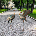 Two decorative copper crane statues with a weathered patina finish in a garden setting.