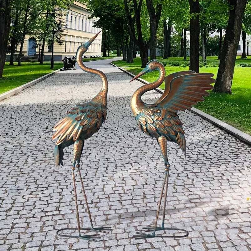 Two decorative copper crane statues with a weathered patina finish in a garden setting.