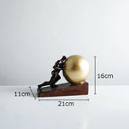 Bronze-colored metal figure statue of a man pushing a golden sphere on a dark wooden base for modern style home decor