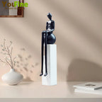 Black glossy luxury metal small figurine of a seated humanoid with elongated limbs on a white pedestal