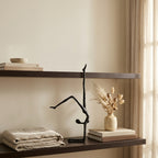 A minimalist black metal abstract sculpture of a dancer, styled on a dark floating shelf next to a small beige ceramic vase with dried grasses and stacked books.