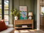 A framed painting of a heron in a light wood console table in a styled living room. The room features dark green paneled walls, a beige sofa with an orange pillow, a white ceramic vase with green foliage, and a white table lamp.