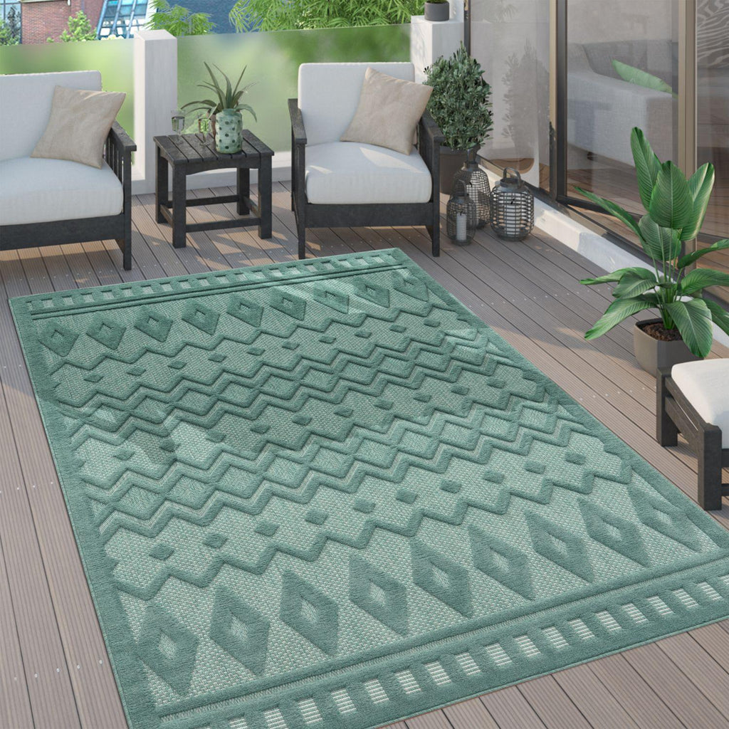 Turquoise geometric trellis pattern indoor outdoor rug on a wooden patio deck with white lounge chairs.