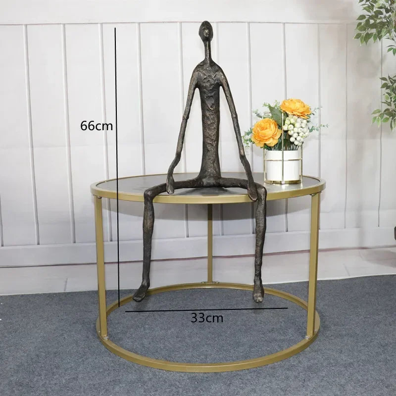 Large abstract sitting human sculpture in dark bronze with elongated limbs on a circular gold metal table