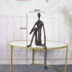 Large abstract sitting human sculpture in dark metal with elongated limbs on round table modern figurine design