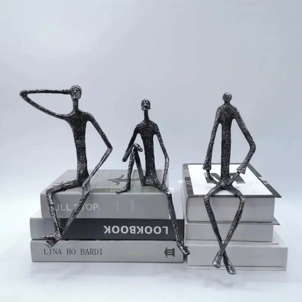 Large abstract sitting human sculpture featuring three slender textured metallic figurines atop stacked books