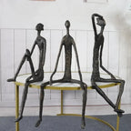Large abstract sitting human sculpture featuring three bronze-colored metal figurines seated on a round table