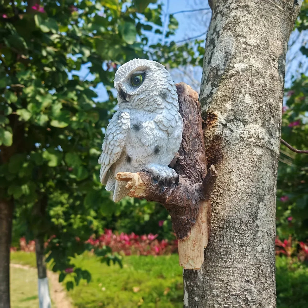 Realistic white owl statue perched on a branch mounted to a tree trunk in a garden.