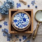 A stack of round ceramic plates with a blue floral and botanical pattern on a white background, presented inside a square wooden box on a linen surface with blue flowers, a magnifying glass, and small blue beads as styled props. [[SKU:HOA-MOR-KIT-007]]