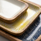 A close-up of stacked rectangular ceramic dishes with rounded wooden rims. The top dish is plain white, the middle dish has a yellow sunburst pattern on a white base, and the bottom dish features a navy blue and white chevron pattern.