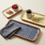 Three rectangular serving trays with light wood rims are arranged on a beige fabric surface. The trays have different printed patterns: one white with a yellow sunburst, one blue and white chevron, and one white with a yellow sunburst.