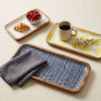 Three rectangular serving trays with light wood rims are arranged on a beige fabric surface. The trays have different printed patterns: one white with a yellow sunburst, one blue and white chevron, and one white with a yellow sunburst. [[SKU:HOA-MOR-KIT-028]]