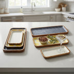 A set of rectangular ceramic serving trays with warm wood-look rims, displayed on a white kitchen countertop with a blurred kitchen background. The trays feature cream, yellow, and textured dark gray glazes.