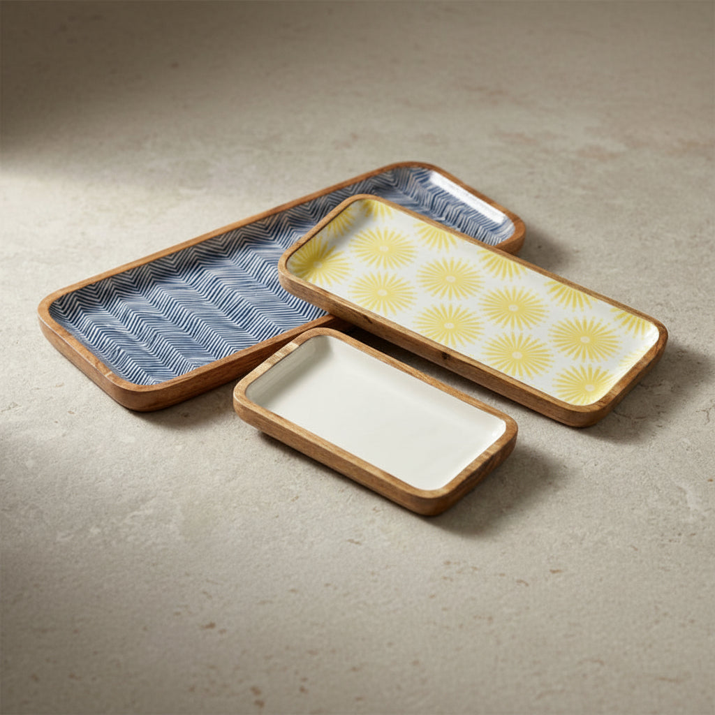 Three rectangular ceramic dishes with light wood rims arranged on a textured beige surface. One dish has a blue and white geometric pattern, one has a yellow sunburst pattern on a white background, and one is plain white.