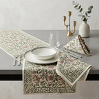 A styled dining table setting featuring a cream table runner and matching napkins with a pink and green floral and botanical pattern, set on a gray table with white plates, cutlery, a glass, and a gold candelabra.