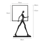 Black metal sculpture of a stylized human figure holding a square frame next to a comforting velvet sofa cushion for living room decor