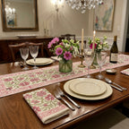 A styled dining table set for a dinner party with a floral table runner, matching napkins, cream plates, crystal glasses, candles, and a wine bottle in a warmly lit dining room.