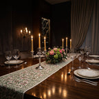 A long floral table runner with pink and green botanical patterns laid across a dark wooden dining table set with white plates, wine glasses, silver cutlery, and lit taper candles.