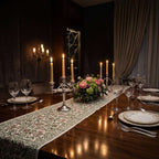 A long floral table runner with pink and green botanical patterns laid across a dark wooden dining table set with white plates, wine glasses, silver cutlery, and lit taper candles. [[SKU:HOA-TR-EGPK-009]]