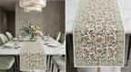 A decorative floral table runner shown in a dining room setting and in a close-up detail view. The runner has a cream background with a dense pattern of pink flowers and green leaves, bordered by ornate matching motifs.