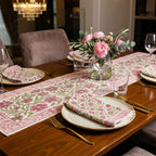 A floral table runner and matching folded napkins with gold flatware are arranged on a wooden dining table, alongside a glass vase of pink flowers, plates, and wine glasses in a styled dining room setting.