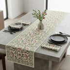 A floral table runner and matching napkins styled on a concrete dining table with black plates, cutlery, wine glasses, and a small vase of pink flowers in a bright room.