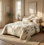 A beige quilted comforter with a gray bird pattern, styled on a bed in a warm, sunlit bedroom with neutral decor.