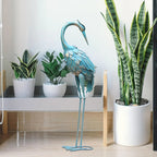 Blue crane metal garden statue with a blue patina finish standing next to potted snake plants.