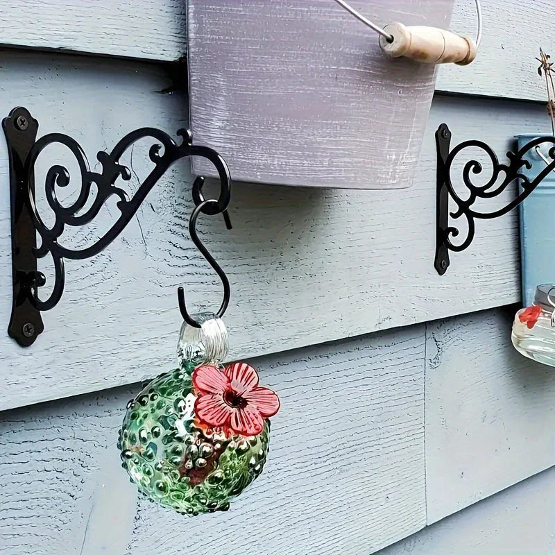 Two black metal decorative swirl wall hooks mounted on a wooden wall holding a bucket and glass ornament. [[SKU:HOA-GDN-003]]