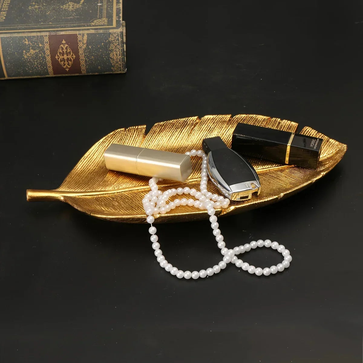 Gold leaf shaped decorative tray holding pearls and lipstick on a dark surface