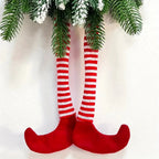 Close-up of dangling legs in red and white striped fabric socks with red pointed elf shoes hanging beneath frosted pine branches against a white wall.