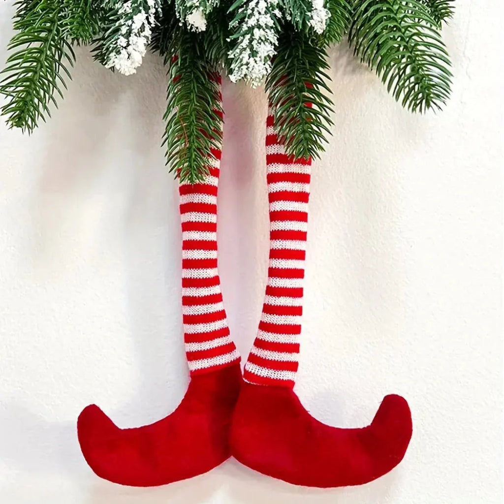 Close-up of dangling legs in red and white striped fabric socks with red pointed elf shoes hanging beneath frosted pine branches against a white wall.