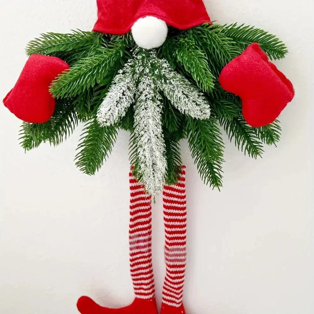 Close-up of a gnome hanging ornament showing a red hat, green pine foliage, soft fabric mittens, and long striped legs against a white wall.