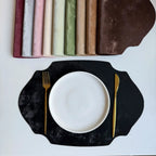 Black scalloped placemat with a white plate, gold fork, and knife on a light gray background.